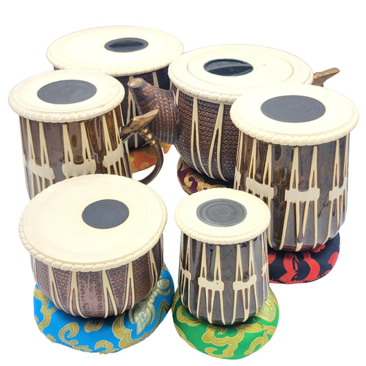 Various ceramic tabla vessels, with glazes that resemble woodgrain and copper, cream-colored drumheads, and colorful cushioned bases, arranged together on a plain light background